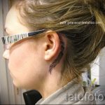 Фото тату тонкой ветви дерева за ухом, выполненной в черном цвете. Элегантный и минималистичный рисунок татуировки.