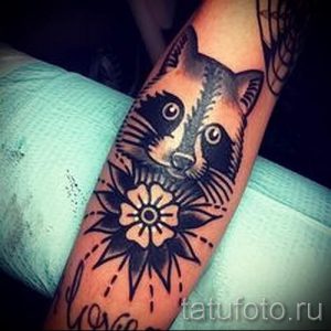 Фото тату енота с цветком в стиле олдскул на руке. Черно-серый рисунок с четкими контурами и традиционными элементами.