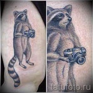 Фото тату енота с фотоаппаратом на руке. Рисунок выполнен в реалистичном стиле с использованием черно-серых оттенков.