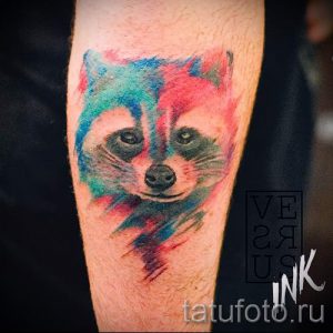 Акварельная татуировка енота на руке, выполненная в ярких и насыщенных цветах, с эффектом размытия и брызг краски.