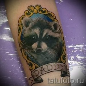 Реалистичная татуировка енота, заключенного в золотую овальную рамку, под которой расположена надпись "DADDY" на ленте.
