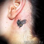Фото тату маленькой пчелы за ухом, выполненной в реалистичном стиле с использованием черных и коричневых оттенков. Изящный рисунок татуировки.