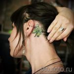 Фото татуировки с изображением четырехлистного клевера, украшенного маленькими цветами, на затылке девушки. Яркий и красочный дизайн.