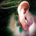 Фото татуировки с тремя маленькими зелеными четырехлистными клеверами, расположенными вдоль линии роста волос за ухом. Изящный и минималистичный дизайн.