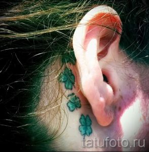 Фото татуировки с тремя маленькими зелеными четырехлистными клеверами, расположенными вдоль линии роста волос за ухом. Изящный и минималистичный дизайн.