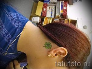 Фотография простой татуировки клевера на затылке, выполненной в зеленом цвете. Небольшой и аккуратный рисунок татуировки.