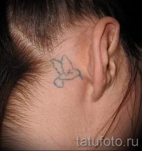 Фотография простой татуировки птицы, выполненной в сером цвете на затылке. Небольшой рисунок татуировки для тех, кто ценит лаконичность.