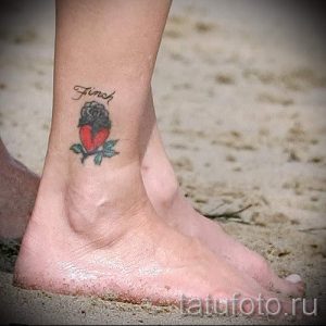 Фото тату сердца, обвитого цветами, на лодыжке. Рисунок татуировки выполнен в традиционном стиле с использованием ярких цветов и четких контуров.