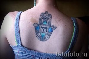 Фото татуировки Хамса на спине девушки, с использованием бирюзовых и серых оттенков. Рисунок татуировки с детально проработанными узорами и надписью.