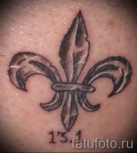 Фото тату: маленькая лилия (флёр-де-лис) в черно-серых тонах с числом '131'. Простой и лаконичный рисунок татуировки.