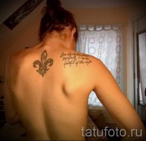 Фото тату на спине: лилия (флёр-де-лис) в черно-серых тонах с цитатой на латыни. Элегантный рисунок татуировки.