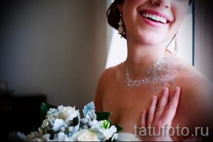 Фотография девушки с блестящей татуировкой, имитирующей ожерелье, на шее. Она улыбается и держит букет цветов.