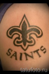 Фото тату: черно-белый рисунок флер-де-лис с надписью "SAINTS" на плече. Лаконичный и стильный дизайн.