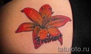Фото тату: оранжевая лилия с надписью "Freiheit" на плече. Оригинальный рисунок татуировки, сочетающий в себе цветочный мотив и текст.