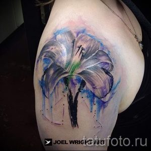 Фото тату лилии в акварельном стиле на плече. Нежные переходы цветов, размытые контуры и легкий эффект брызг.