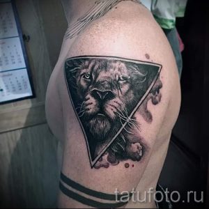 Фото тату: треугольник с изображением головы льва, расположенный на плече. Реалистичный рисунок татуировки в черно-белом стиле с использованием теней.