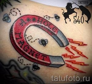 Фото тату подковы с оранжевыми молниями, мухами и надписью "A HOLE MAGNET" на руке. Стильный рисунок татуировки в стиле олдскул с использованием ярких цветов.