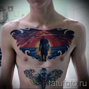 Фото тату с изображением человека, стоящего на берегу моря на фоне заката, выполненного на груди. Татуировка выглядит драматично и символично.