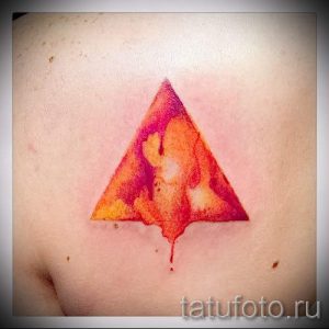Фото акварельной татуировки на груди с изображением треугольника, выполненного в ярких оранжевых и красных тонах. Эффект растекающейся краски.