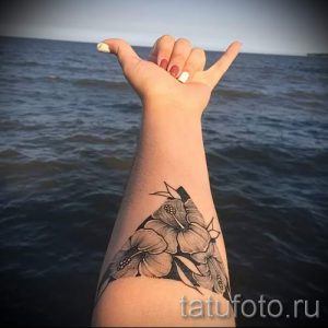 Фото татуировки на предплечье, изображающей несколько цветков гибискуса. Рисунок выполнен в черно-серых тонах с высокой детализацией.