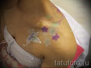 Фотография блестящей татуировки на плече, изображающей цветы и бабочку, выполненной с использованием серебристых и фиолетовых блесток.