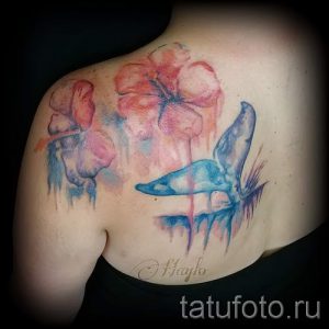 Фото тату акварельного кита и цветов на лопатке. Рисунок выполнен в нежных розовых и голубых тонах с эффектом размытия и брызг краски.