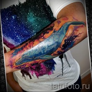 Фото тату: красочная акварельная татуировка кита, плывущего в космическом пейзаже с галактиками и звездами на руке.