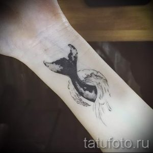 Фото тату: минималистичный рисунок хвоста кита на женском запястье, выполненный в технике тонких линий и черного цвета.