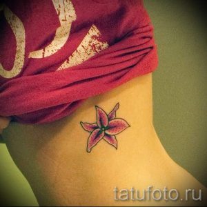 Фото тату маленького цветка лилии на женской шее. Изящный рисунок татуировки выполнен в нежных розовых и фиолетовых тонах.