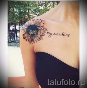 Фото тату подсолнуха с надписью "You are my sunshine" на плече. Черно-серый рисунок татуировки с реалистичным изображением цветка и каллиграфической надписью.