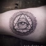 Черно-белое фото татуировки на предплечье с изображением всевидящего глаза в треугольнике, окруженного облаками и лучами света. Детализированный рисунок татуировки.