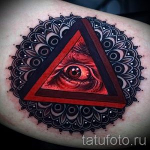 Цветное фото тату на предплечье с изображением красного глаза в треугольнике, окруженного сложной мандалой. Яркие цвета и детализированный рисунок.