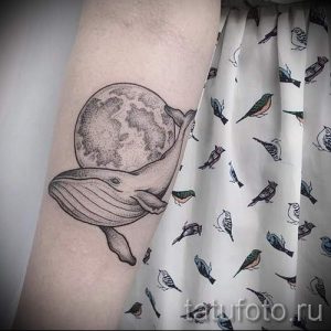 Фото минималистичной татуировки кита под луной, окруженного стаей птиц на предплечье. Черно-белый рисунок с тонкими линиями.