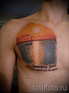 Фото тату с изображением морского пейзажа на груди, включающего закат, парусник и надпись с датами. Татуировка выполнена в реалистичной технике.