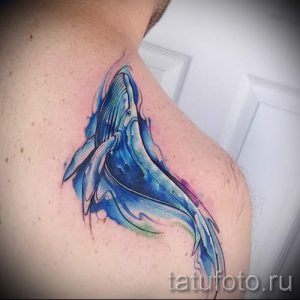 Фото тату кита в акварельном стиле на плече. Цветная татуировка с использованием мягких переходов цветов и размытых контуров. Нежный и романтичный дизайн.