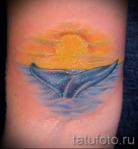Фото тату китового хвоста на фоне заката. Акварельная татуировка с использованием мягких переходов цветов и размытых контуров. Романтичный и нежный дизайн.