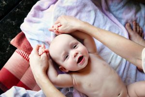 Фотография счастливого младенца, лежащего на руках у родителей. Младенец улыбается и тянется ручками к камере. Теплые и уютные цвета.