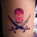 Фото тату с изображением пиратского черепа и скрещенных костей, выполненного блестками на коже. Цвета: розовый, золотой, синий.