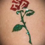 Фото тату на коже: красная роза с зелеными листьями, выполненная с использованием глиттера. Яркий и необычный рисунок татуировки.