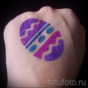 Фотография татуировки в виде разбитого сердца, выполненной розовыми и фиолетовыми блестками на руке. Оригинальный и привлекательный рисунок.