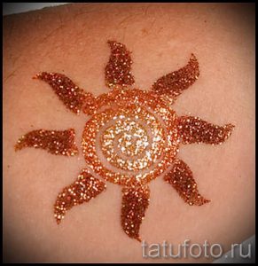 Фотография блестящей татуировки в виде солнца, выполненной золотыми блестками на коже. Яркий и привлекательный рисунок.