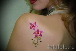 Фотография татуировки в виде двух звезд и стебля с листьями, выполненной розовыми блестками на плече. Нежный и привлекательный рисунок.