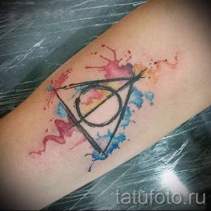 Фото тату: Акварельная татуировка Дара Златой Канарейки (Deathly Hallows) из Гарри Поттера на предплечье. Яркие цвета и размытые линии.