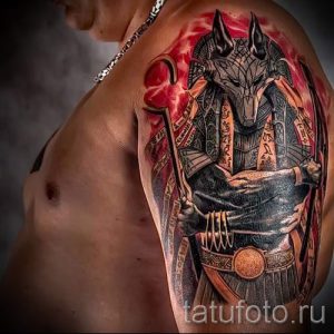 Фото тату Анубиса на плече. Изображение выполнено в ярких цветах с использованием реалистичных деталей. Анубис изображен в полный рост, с символами и иероглифами.