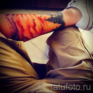 Фото тату на руке: пейзаж с сосновым лесом, переходящим в закатное небо. Использованы оранжевые, красные и черные цвета.