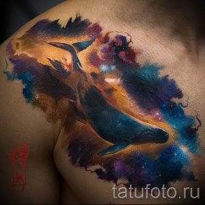 Фото татуировки на плече с изображением кита, плывущего в галактике. Рисунок выполнен в технике акварели с использованием ярких и насыщенных цветов.