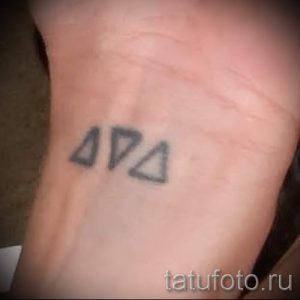 Черно-белое фото татуировки на запястье, изображающей три небольших треугольника, расположенных рядом друг с другом.