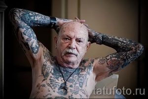 Фото тату пожилого мужчины, практически полностью покрытого татуировками в различных стилях. Мужчина стоит, заложив руки за голову.