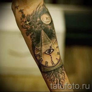 Татуировка на предплечье с изображением пирамиды, Всевидящего Ока и часов. Рисунок татуировки выполнен в реалистичном стиле с использованием черных и серых оттенков.