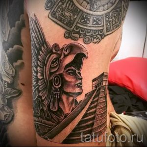 Черно-белая татуировка на руке с изображением воина в головном уборе, напоминающем ацтекский, на фоне пирамиды.